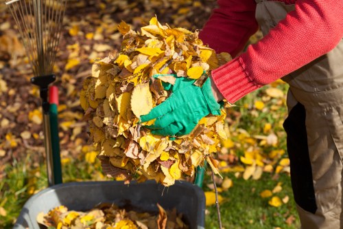 A person wearing red long-sleeved shirt and bright green gloves is lifting a large bundle of yellow and brown autumn leaves. The bundle appears freshly gathered and is being placed into a grey rubbish collection or yard waste bin located outdoors on a grassy area. In the background, more fallen leaves cover the ground, and a garden rake with a black handle and orange head is visible leaning against a shrub or garden border. The scene suggests typical autumn leaf clearance, which may be undertaken by a professional waste removal service such as Office Clearance Stratford, operating in Stratford or nearby areas. The natural outdoor setting features leafy surroundings consistent with a residential garden or park environment, illustrating waste management activities related to seasonal clearing tasks.