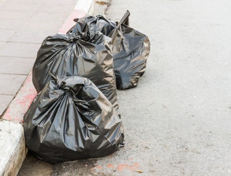 Two black plastic rubbish bags filled with waste are situated on a paved footpath next to a concrete curb, with a small metal rake leaning against the bags. The bags appear to be tightly closed, and the surface of the plastic shows some creases and folds, indicating they are filled with rubbish. The ground surface is smooth and grey, and the curb is made of weathered concrete, separating the footpath from the road. The scene is outdoors, with natural lighting and no visible background details beyond the immediate area around the rubbish bags. This image relates to waste collection or rubbish removal services typically offered by companies like Office Clearance Stratford, serving areas within Stratford or nearby postcodes, emphasizing basic waste management needs in an urban environment.