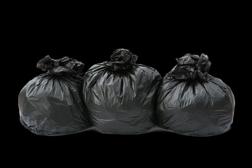 Three sealed black rubbish bags made of plastic are positioned side by side on a dark background, with the middle bag slightly in front of the other two. The bags appear full and are knotted at the top, their surface showing a glossy, crinkled texture. The illumination highlights the bags' reflective surfaces and various creases, emphasizing their full capacity. The setting is neutral and uncluttered, focusing solely on the bags without additional objects or context, suggesting they are prepared for removal or disposal. This scene could be representative of waste collection or rubbish disposal in a residential or commercial environment, aligning with services offered by Office Clearance Stratford. The image's simplicity and clear visual details under normal lighting conditions support it being useful for illustrating rubbish removal tasks within the Stratford area, reflecting typical waste items ready for collection.