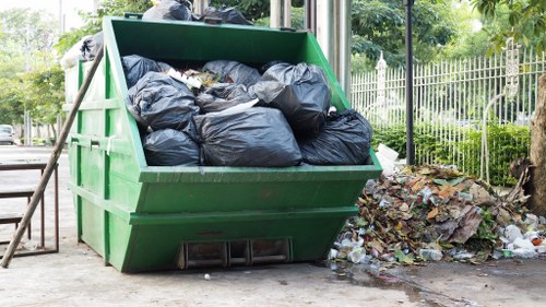 A large green skip filled with tightly packed black rubbish bin bags, positioned outdoors on a paved surface near a metal fence and trees. The skip is situated beside a pile of mixed waste, including paper and plastic, discarded on the ground. In the background, there are trees and a green hedge, indicating a residential or park nearby. The scene is captured in natural daylight, with the skip appearing used for waste collection as part of a rubbish removal service, with the environment suggesting an urban or suburban setting that may be serviced by companies like Office Clearance Stratford, based in the Stratford area. The arrangement emphasizes the practical aspect of waste disposal and clearance within a local community, with the surrounding environment reflecting typical city or town landscape in the UK.