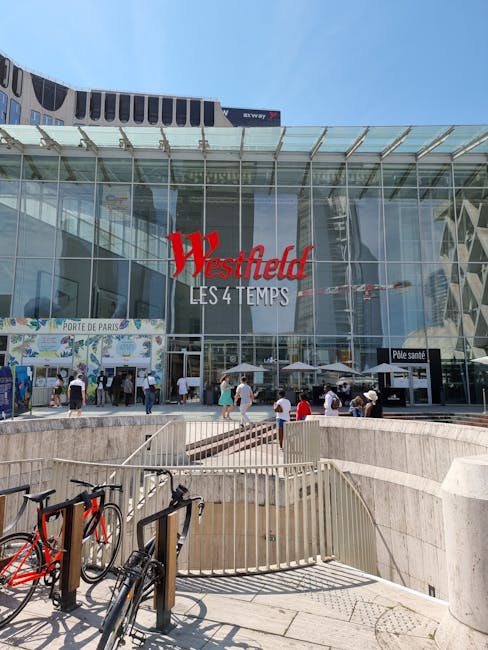 The image depicts the modern glass facade of Westfield, a large shopping centre in Stratford, with the red and white Westfield sign prominently displayed above the entrance. In front of the building,