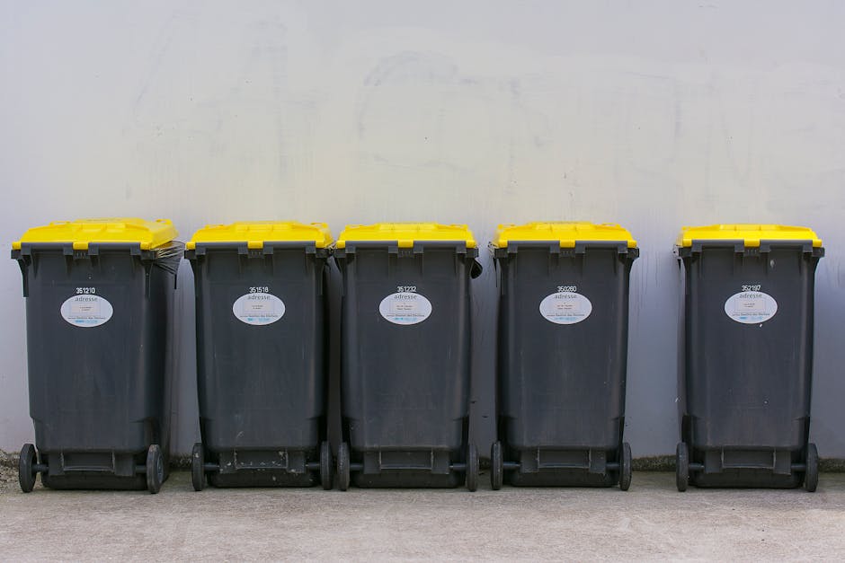 A large green waste collection bin filled with various discarded cardboard boxes, some flattened and others still in their original shape, along with a few white packaging materials and smaller cardbo