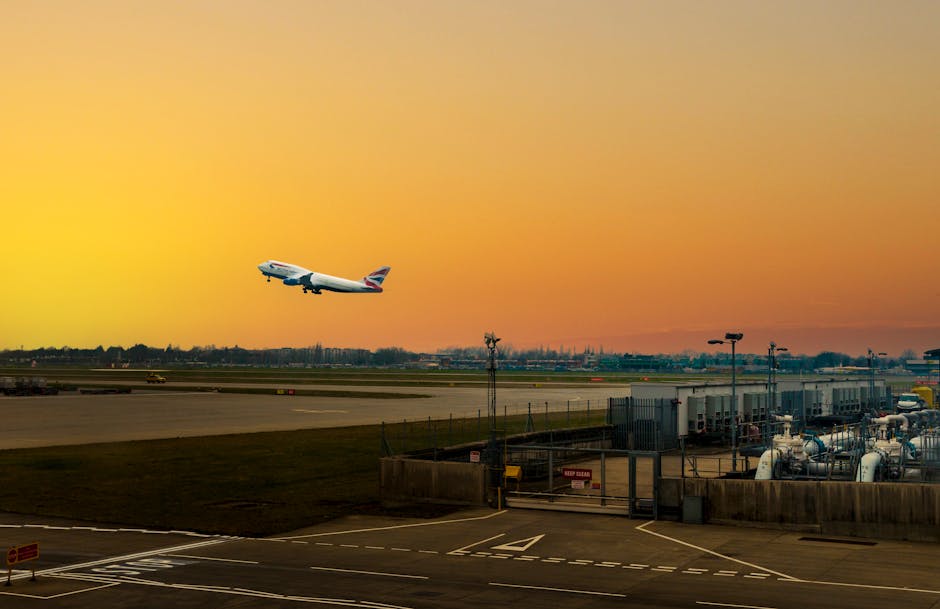 A commercial airplane with a predominantly white fuselage and a red, white, and blue tail fin is ascending into a sky painted with warm orange and yellow hues during sunset, against the backdrop of an