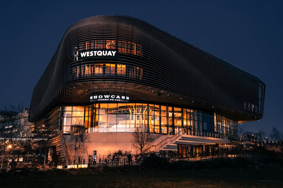 A modern multi-storey building at night with a distinctive curved exterior, featuring horizontal metal cladding in dark tones and large glass windows illuminated from within. The upper floors display 